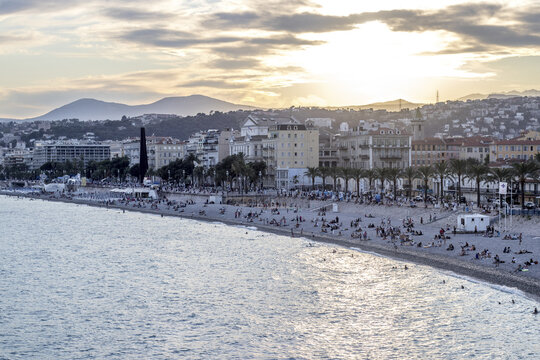 View of the beach filled with people enjoying the sun and sea, contrasted against the city skyline and distant mountains, Nice, France.
