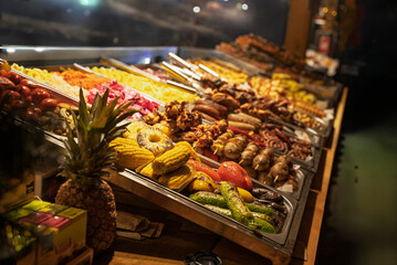Vibrant street food stall at a night Christmas market featuring grilled meat skewers, sausages, corn on the cob, and colorful vegetables under warm bokeh lights. A cozy festive atmosphere