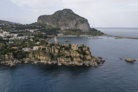 Aerial view of the rugged coastline meeting the shimmering sea, dominated by the Rocca's imposing presence and the ancient towers standing guard, Cefalu, Sicily, Italy.