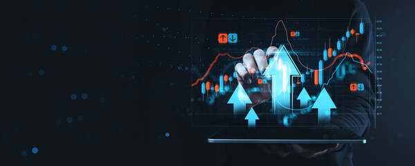 Futuristic concept of rising stock market with candlestick chart and arrows on digital interface background held by hand on tablet device screen.