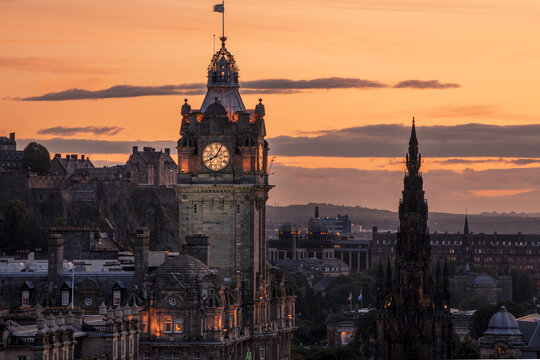 View of the Balmoral clock tower illuminated under the pastel sky, with the Scott Monument standing tall in the cityscape, Edinburgh, Scotland, United Kingdom.