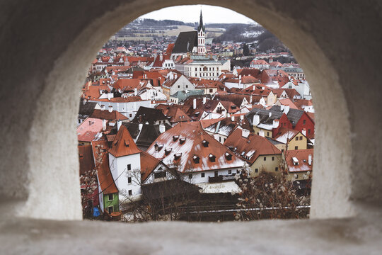 View of a medieval townscape unfolds through a stone arch, the terracotta rooftops dusted with snow, contrasting with the overcast sky, Cesky Krumlov, South Bohemian Region, Czechia.