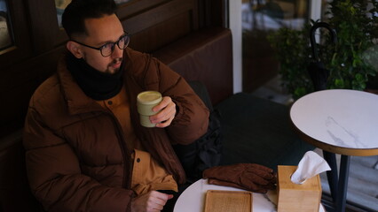 Young man in a jacket sits on an open cafe terrace on an autumn day drinking matcha latte