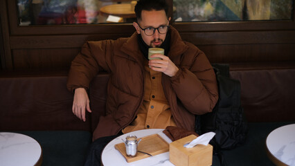 Young man in a jacket sits on an open cafe terrace on an autumn day drinking matcha latte
