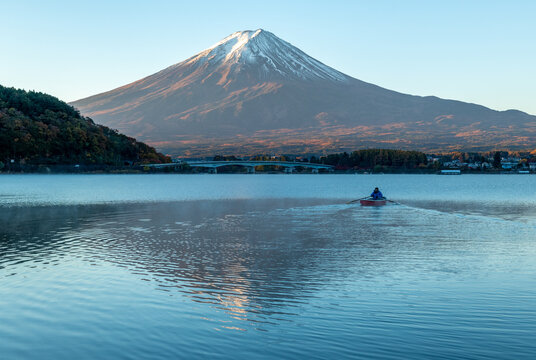 View of Mount Fuji's snow-capped peak mirrored in the tranquil lake as a lone rower glides through the serene waters, Fujikawaguchiko, Yamanashi, Japan.