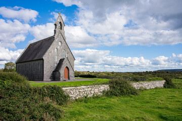 An Irish little church with an orange door surrounded by a little stone wall and a green field with bushes. The blue sky is clouded
