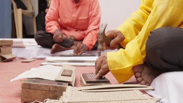 Two artisans sit and make Jacquard punch cards by hand using hammers and punches. These cards guide looms to weave detailed silk saree patterns in traditional Indian textiles 