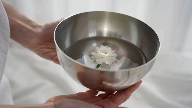 Cinematic close-up of calm human hands holding a shallow brushed metal bowl filled clear water single elegant white flower floating gently inside ripples forming, minimalist zen aesthetic, luxury well