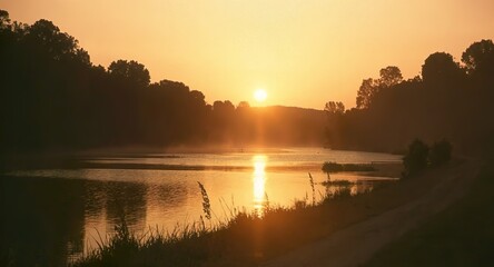 Obraz premium Peaceful sunrise over a calm river with warm golden morning light reflecting on the water.