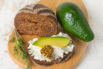 A snack board features ricotta cheese with a slice of avocado and grainy mustard on whole grain rye bread. Healthy sandwiches and ingredients.