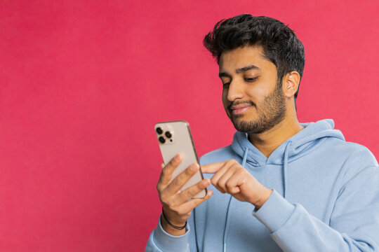 Happy Indian young man browsing social media, smiling while looking at smartphone, enjoying interesting content and typing a message. Relaxed Arabian guy engaging online, isolated on red background - Powered by Adobe