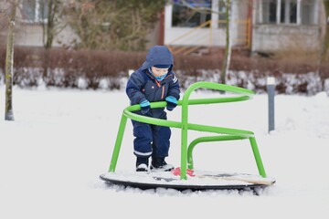 Toddler boy standing on playground carousel in snowy park. Child wearing warm winter snowsuit exploring balance, coordination and independent movement during outdoor winter play on a cold day.