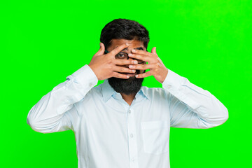 Indian man covers eyes with hands peeks through fingers, imitating a spy with dramatic expression of secrecy. Arabian guy isolated on green chroma key background showing mystery, tension hidden intent