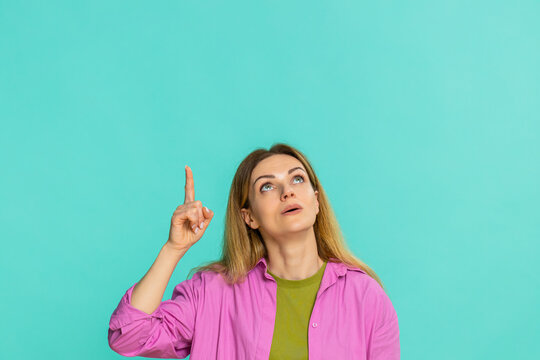 Middle-aged woman points finger upward with serious face, clearly signaling importance, asking to focus attention on something above. Girl isolated on blue background shows emphasis gesture and intent