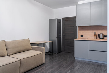 studio apartment design in modern minimalist style with built-in furniture and household appliances. small grey built-in kitchen, wooden countertops,  beige sofa, silver refrigerator