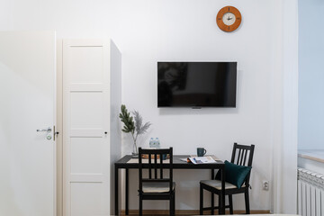 Cozy and functional twin room with TV and workspace. a hotel room or studio with white walls, a large window with curtains, a dark table and chairs, a clock.