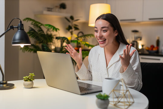 Young woman at night home opens laptop for grant form sees approved message cheers raises hand. Caucasian girl on sofa at table at evening smiles feels joy reads award email jackpot vibe winner mood.