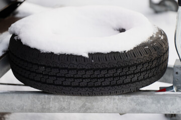 Spare vehicle tire covered with fresh snow resting on metal support frame showing winter storage conditions rubber tread pattern and automotive equipment detail
