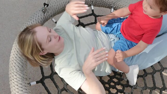 Mother having fun with her little son during a walk. Young mom hugs her baby and swings on a swing