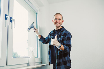Man cleans window with a spray bottle in a bright room during the day while smiling and giving a thumbs up to the camera