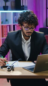 Handsome businessman and his female workmate in traditional hijab sitting in front of laptop in night lighted workroom during video conference with partners and discussing report with charts