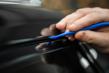A technician uses a blue trim tool to lift a black weatherstrip on a glossy car panel in a luxury dealership service bay. Soft indoor light and shallow depth of field show care.
