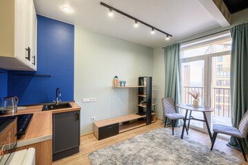Fototapeta premium A compact studio with a modern kitchen, dining area by the balcony, open shelving, wooden furniture, light wood flooring, soft rug, and light green walls. Bright and airy