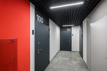 A modern hallway with dark grey doors numbered 100–101, a vibrant red accent wall, grey tiled floor, and black grid ceiling with linear LED lights. Clean, minimalist design.