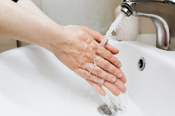Two female hands with white soapy foam in a stream of tap water in the bathroom. The concept of hygiene, cleanliness and protection against coronavirus COVID-19.