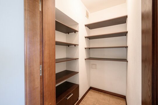 A narrow closet with built-in wooden shelves on two walls, a wooden door, light-colored walls, and a tiled floor. Minimalist design, empty shelves, one drawer unit at the bottom left