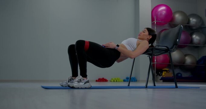 Young woman performs hip thrust exercise on gym mat using a chair and weights in a fitness studio setting