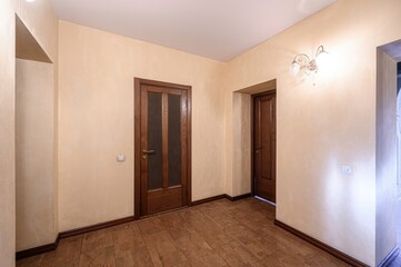 Obraz premium A minimalist hallway with beige textured walls, brown tiled floor, wooden doors (one with frosted glass panels), and wall-mounted sconces providing soft lighting. Clean, uncluttered design