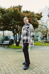 Fototapeta premium Young woman stands in park wearing a checkered coat with fringe, surrounded by trees and benches during a cloudy day