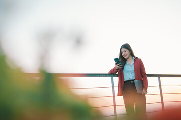 Young professional businesswoman smiling while standing outdoor at sunset, checking messages on her smartphone and holding a laptop, representing modern communication and digital nomad life