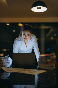 Businesswoman intently focusing on her laptop screen, surrounded by towering stacks of paperwork in a dimly lit office, experiencing stress and pressure to meet an urgent deadline