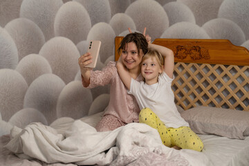 Mother and daughter taking a happy selfie together in bed. Playful moment, family bonding, morning fun with smartphone.
