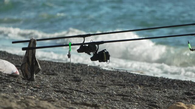 Fishing rods, sea, coastline waiting for a bite as surf fishing poles stand ready on a dark pebble beach under bright sunshine.