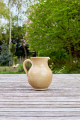 A rustic light brown ceramic pitcher with a handle and spout sits on a weathered wooden table outdoors. Lush green trees and foliage blur in the background, creating a natural, peaceful setting.