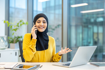 Muslim businesswoman in a hijab and yellow shirt smiling and actively communicating on a smartphone while sitting at a desk with a laptop and tablet in a modern office, enjoying successful work