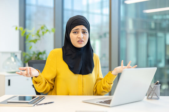 Muslim businesswoman wearing a hijab expressing doubt and frustration while shrugging her shoulders and looking at the camera, sitting at an office desk with a laptop