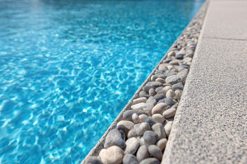 Obraz premium Close-up of pool's edge with pebbles. Clear blue water reflects light. Concrete