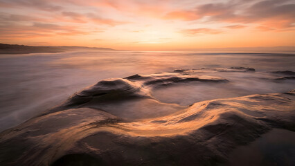 Serene seascape featuring smooth rocky formations along the coastline at sunset, captured with long exposure to create a soft, misty ocean surface and warm golden sky.