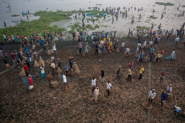 Pabna, Bangladesh - 05 December 2020: Aerial view of workers scattered across the muddy land, their figures like dark smudges against the earth, with vibrant green vegetation along the water's edge.