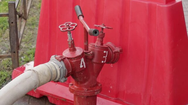 Fire hydrant valve is connected to hose and stands in front of red plastic barrier. Metal handles and fittings are visible.