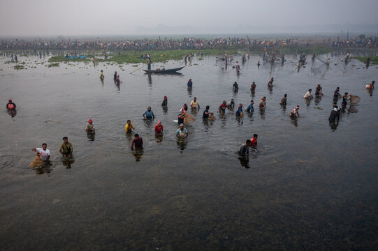 Pabna, Bangladesh - 05 December 2020: Aerial view of a river teeming with figures wading through the murky waters, their forms reflecting the diffuse light under the vast, hazy sky.