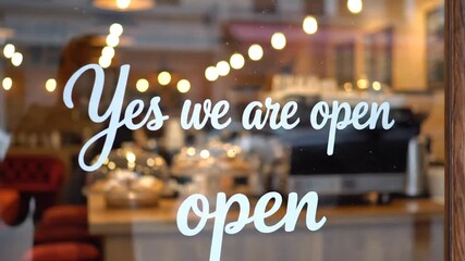 Modern glass entrance door of a boutique shop or cozy cafe displaying a hanging wooden sign with Yes We Are Open text invitation for customers during business hours in a street setting
