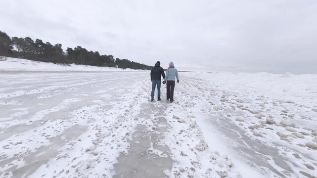Couple holding hands and walking along a snowy winter beach. Peaceful outdoor lifestyle and togetherness concept.