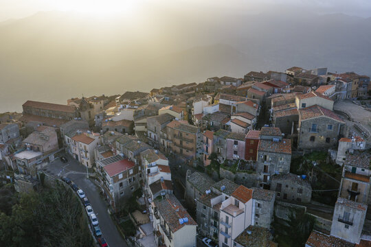 Aerial view of sunlit terracotta rooftops cascade down the hillside towards the sea, an old church standing guard over the ancient town, Pollina, Sicilia, Italy.