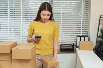 Young asian businesswoman using smartphone verifying package information standing desk surrounded...