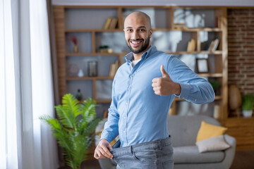 Man smiling and giving thumbs up while pulling out loose jeans to show weight loss success, confident and happy after healthy lifestyle changes at home, feeling fit and motivated